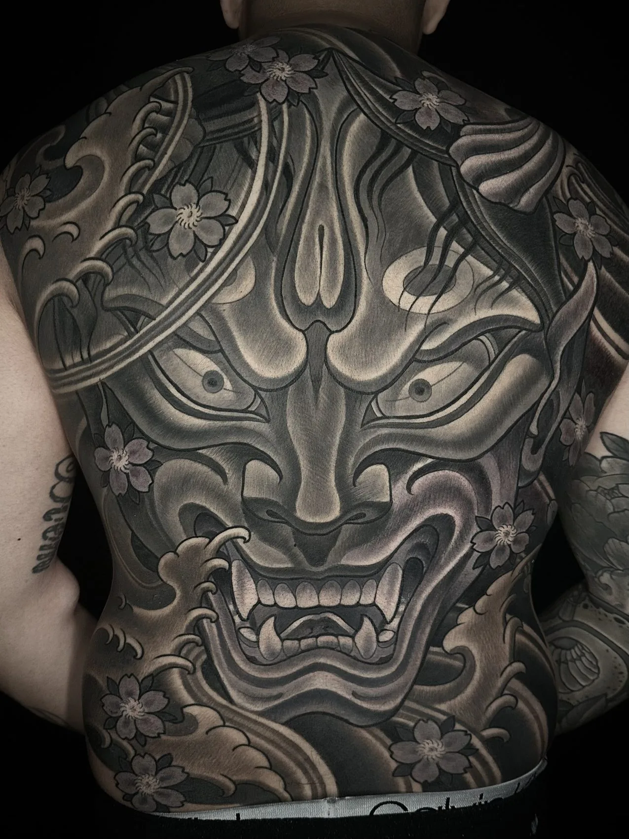 Black and grey full back piece tattoo of a hannya mask with cherry blossoms and waves by Terry Ribera at Remington Tattoo in San Diego.