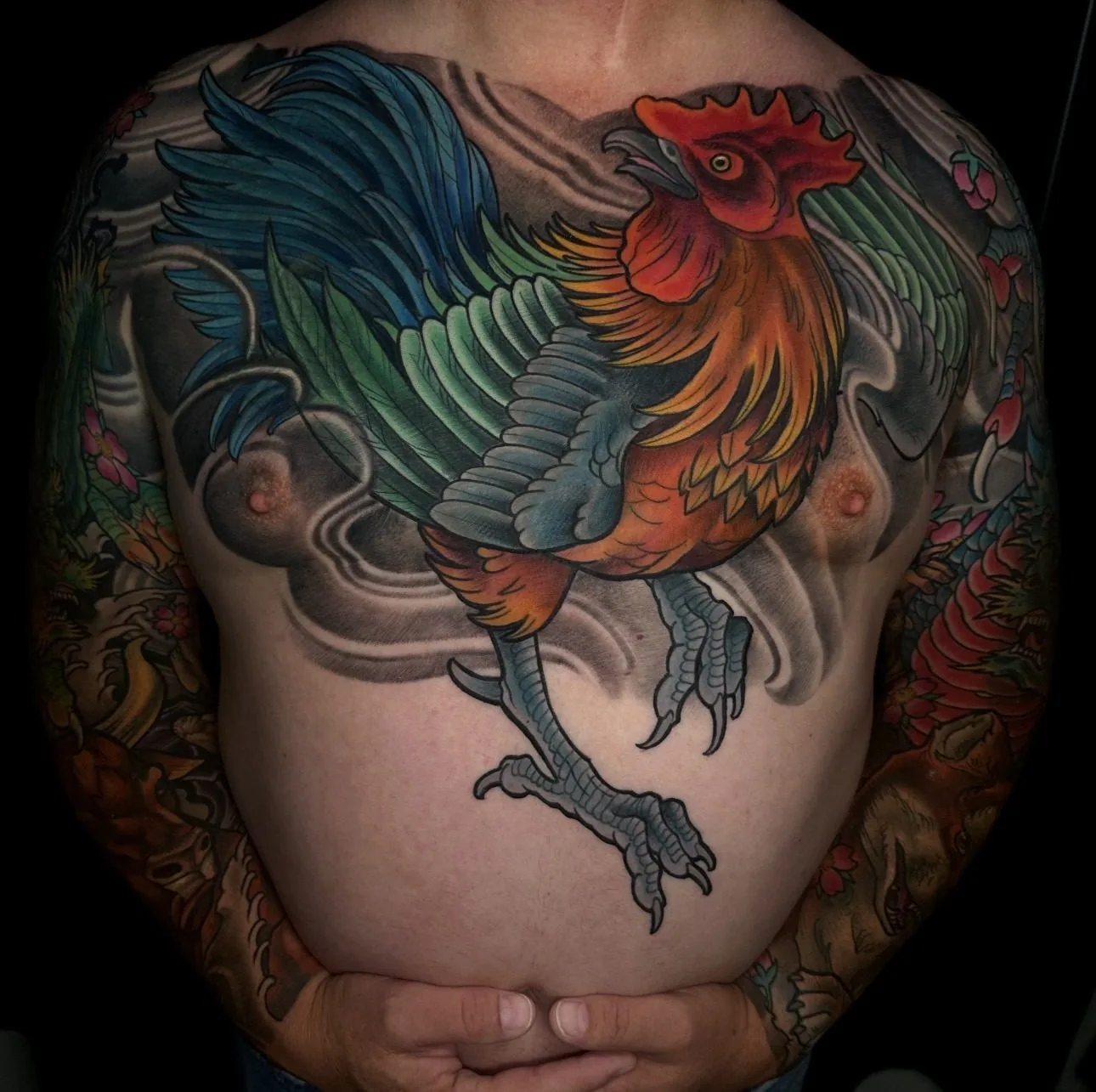 Neo traditional full color chest piece tattoo of a rooster in flight with green and blue tail feathers over black-and-grey wind bars by Terry Ribera at Remington Tattoo in San Diego.